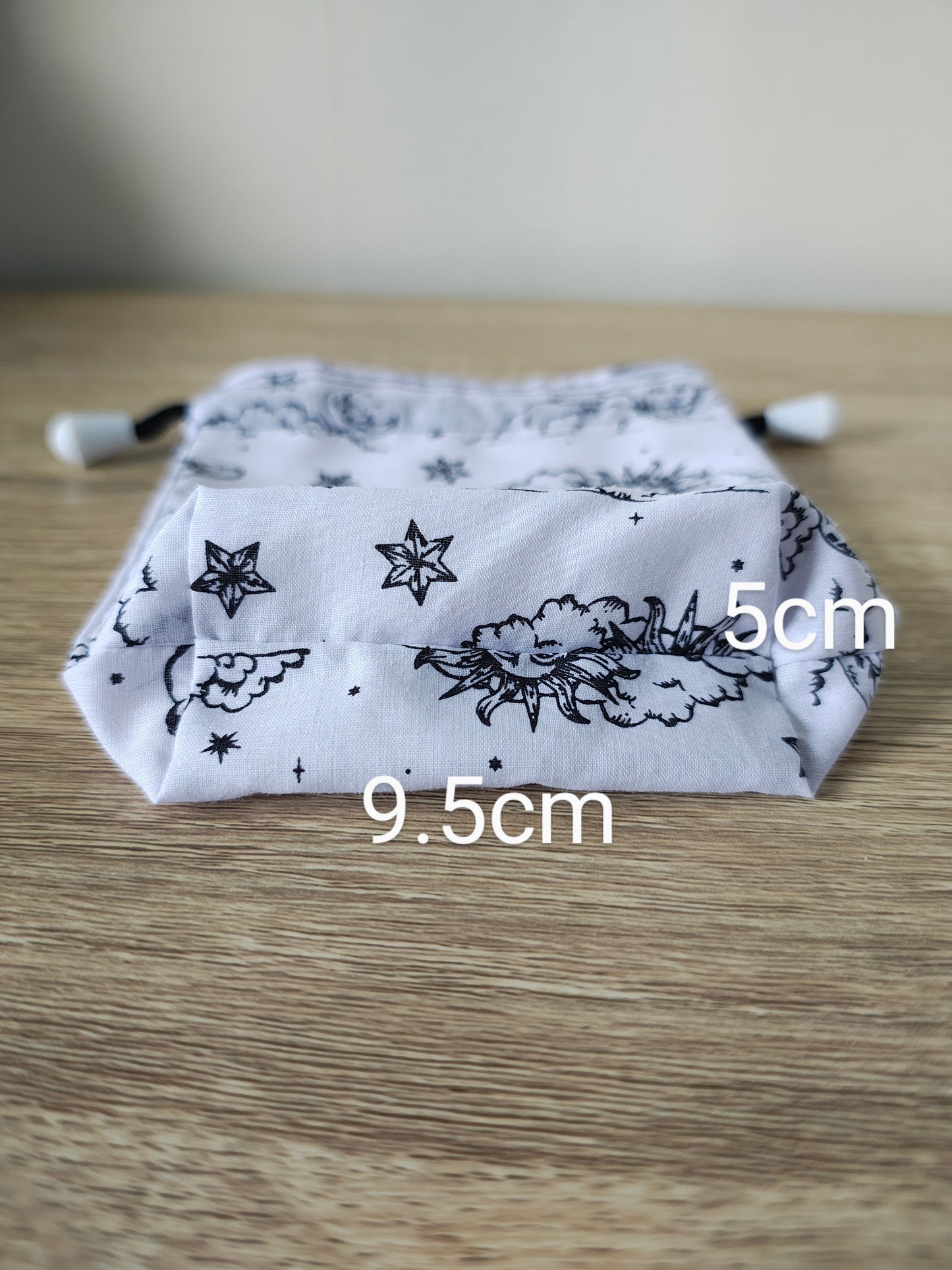 White drawstring bag with black floral and star pattern on a wooden surface, with measurements indicated.