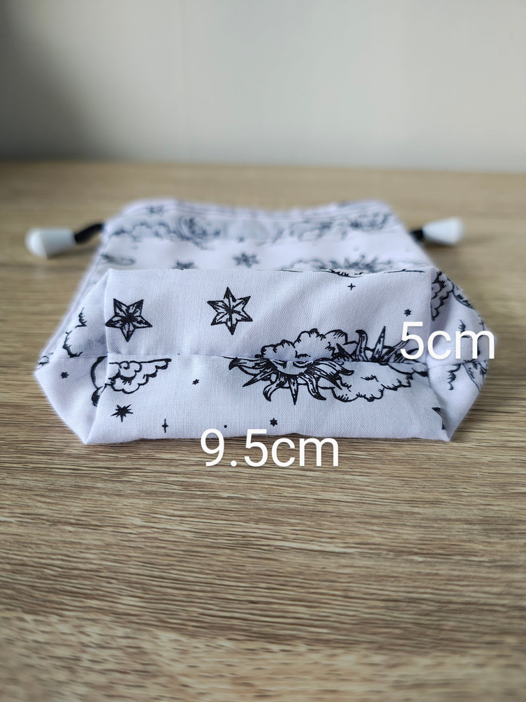 White drawstring bag with black floral and star pattern on a wooden surface, with measurements indicated.