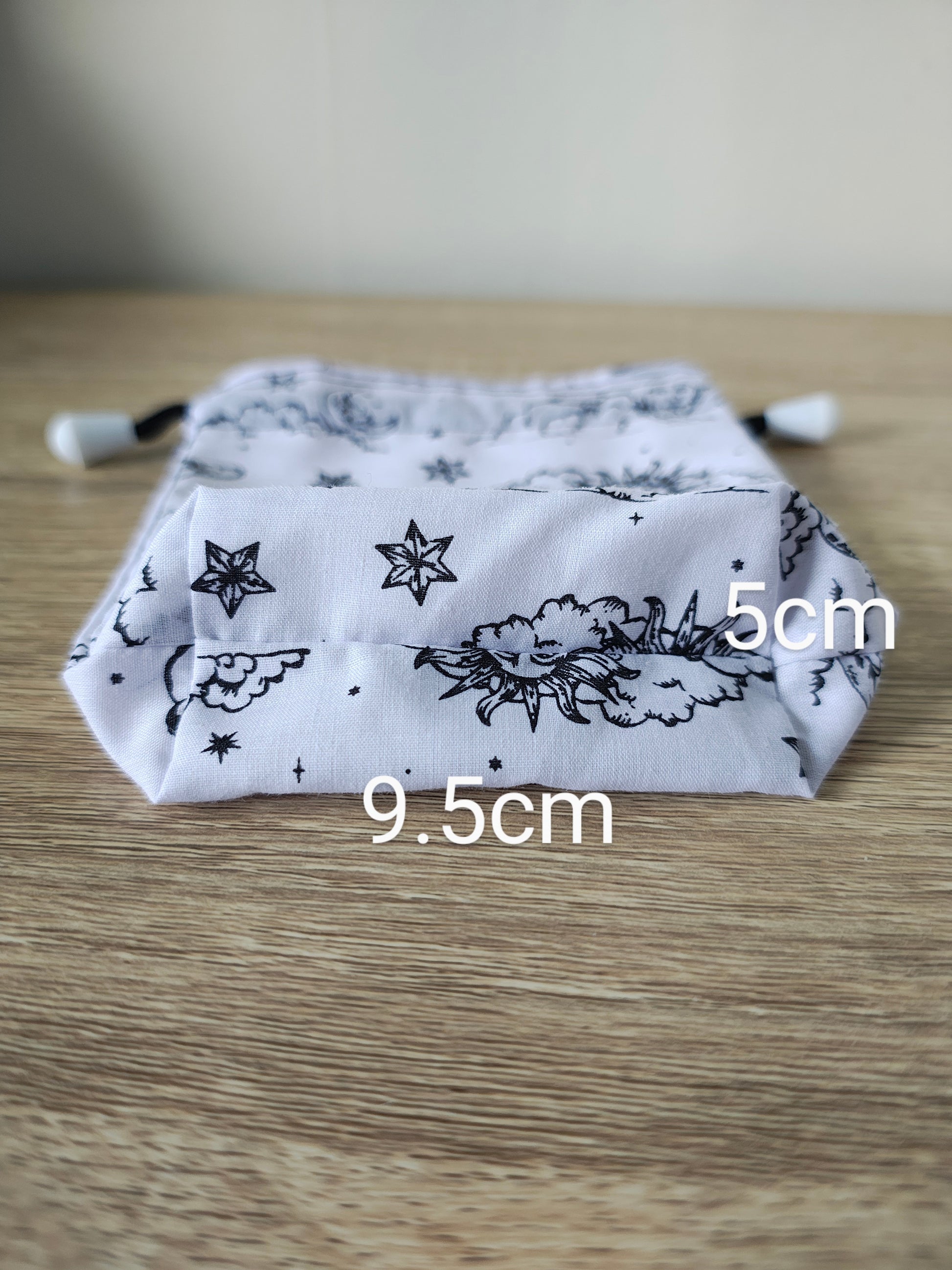 White drawstring bag with black floral and star pattern on a wooden surface, with measurements indicated.