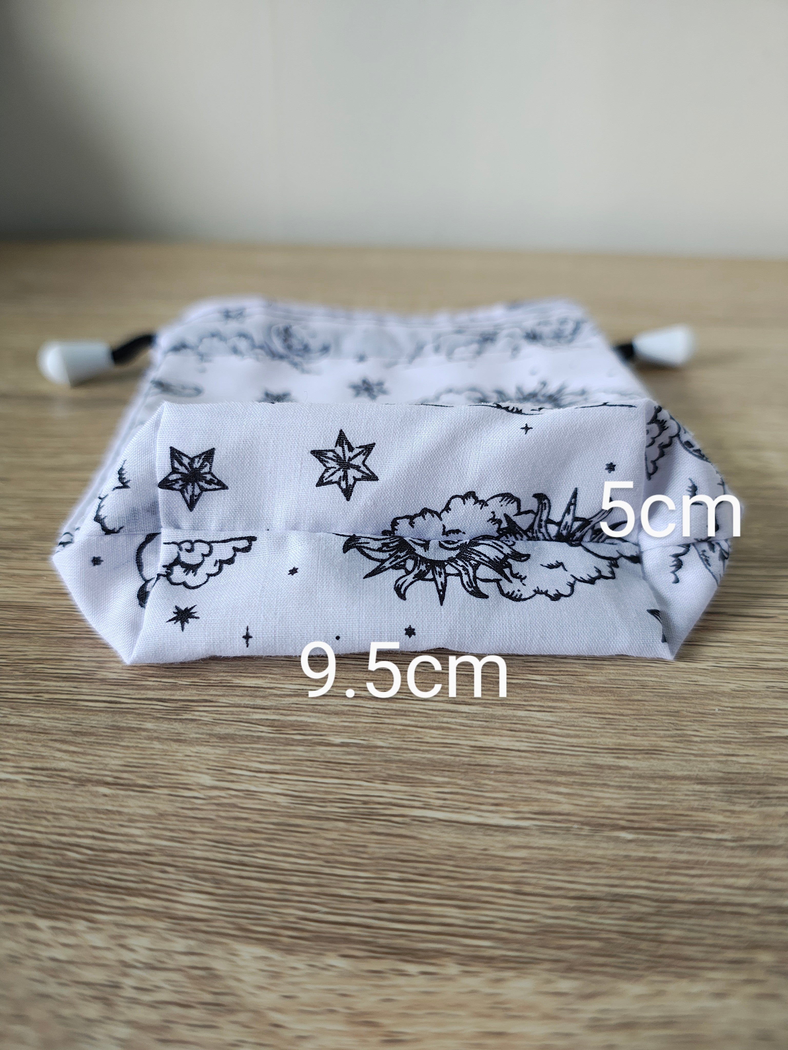 White drawstring bag with black floral and star pattern on a wooden surface, with measurements indicated.