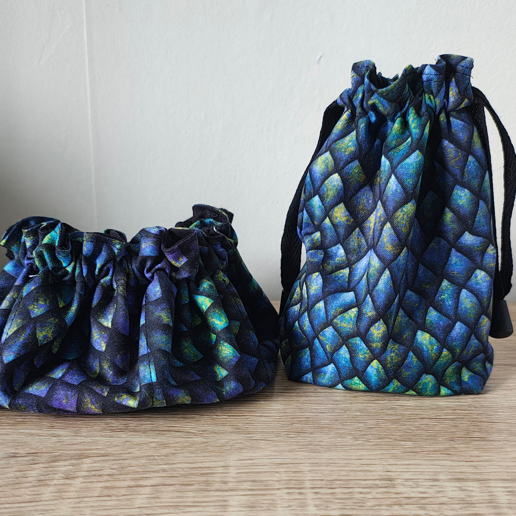 Two drawstring pouches with an iridescent blue and green scale pattern, displayed on a light wood surface against a white wall.