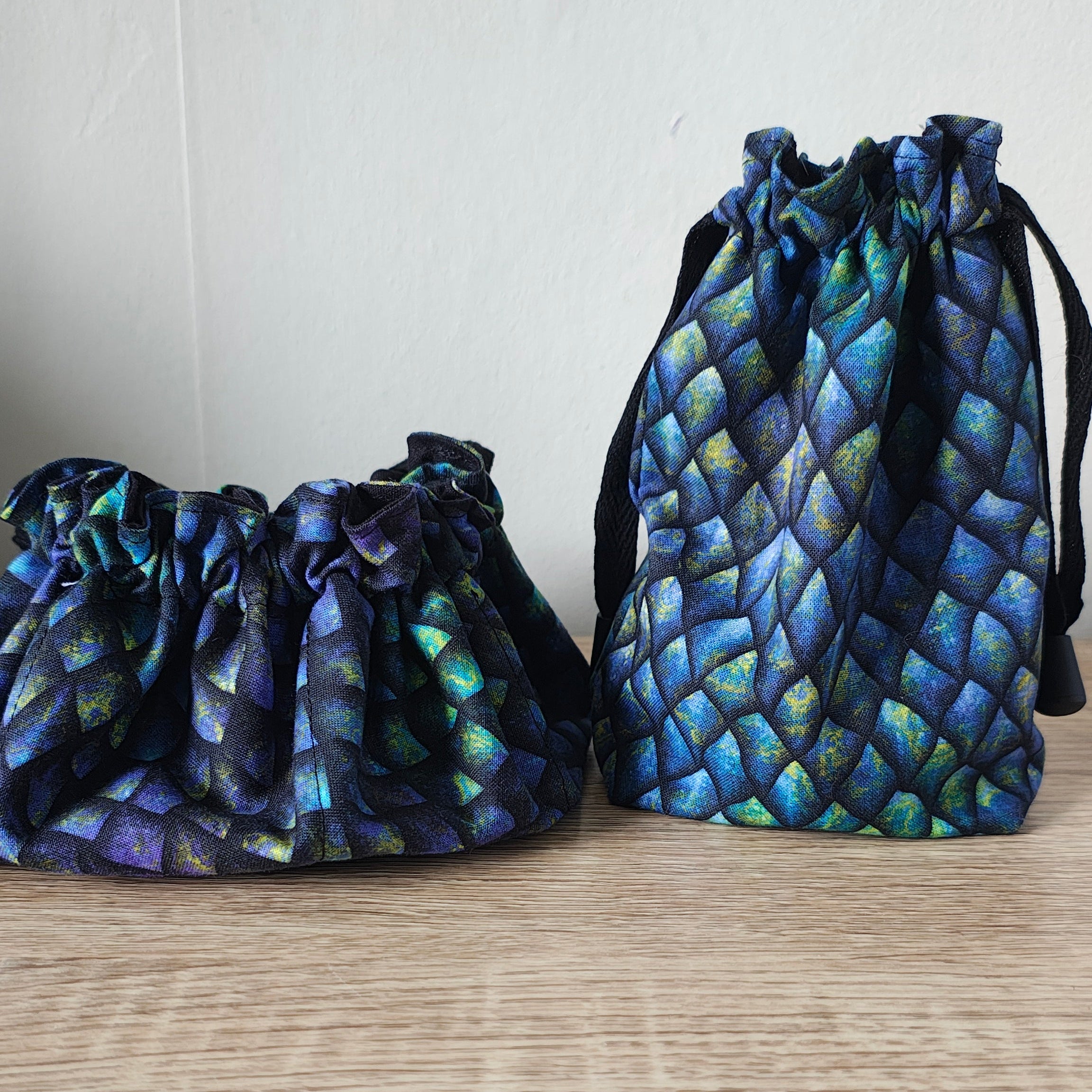 Two drawstring pouches with an iridescent blue and green scale pattern, displayed on a light wood surface against a white wall.